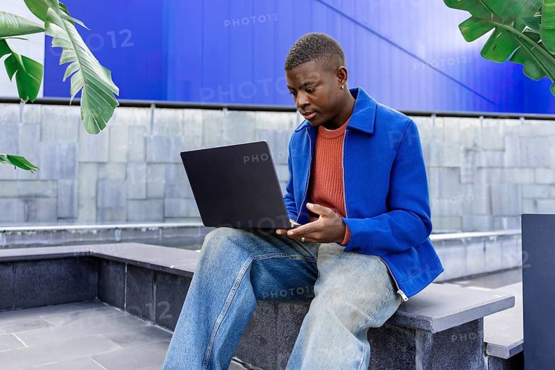 Young African man with short hair sits on a bench, using a laptop in a modern outdoor setting with greenery and a blue wall in the background