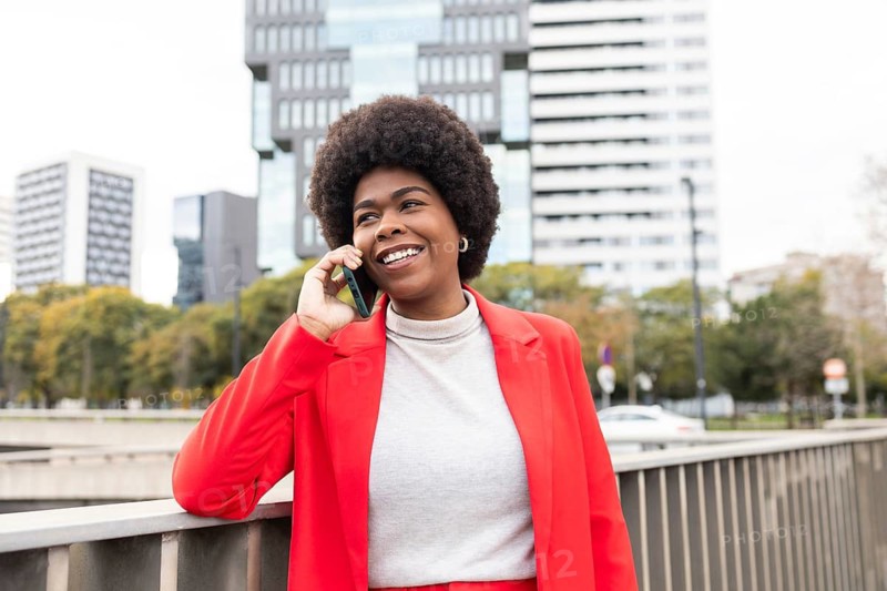 Smiling woman in a vibrant red jacket stands by an urban rail, chatting on her phone. Her confident posture and ethnic style embody a modern urban lifestyle