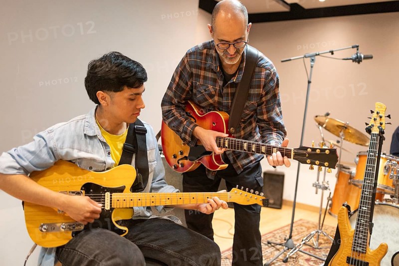 In a recording studio, a guitar instructor guides a student, who is holding a guitar, through a session. Instruments and microphones are set up around them