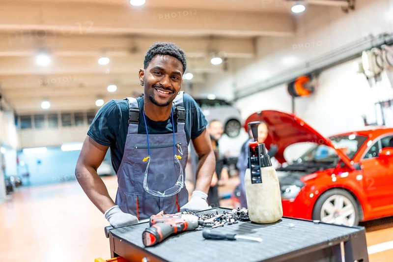 Confident mechanic smiling while posing next to a workbench with tools and equipment in a bustling auto repair shop