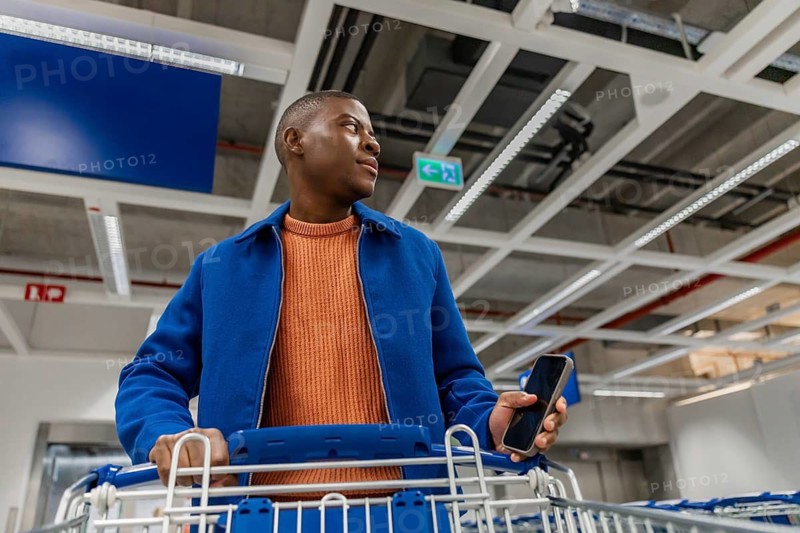 A young man stands in a busy store with a shopping cart. He holds a smartphone in one hand and looks around. The setting has bright lights and shelves in the background