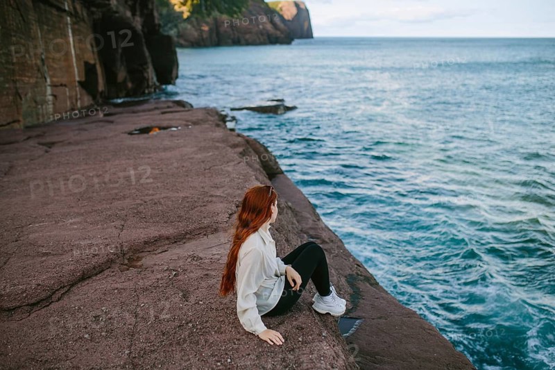 A woman enjoys a serene moment sitting on a rocky ledge overlooking the waves at Flatrock, Newfoundland. Her red hair contrasts beautifully with the blues of the sea