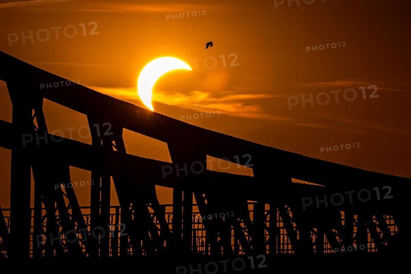 Partial solar eclipse above a large industrial bridge structure, creating a dramatic silhouette against a glowing orange sky at sunset