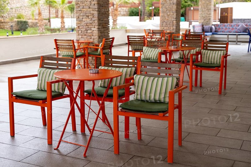 A stylish outdoor seating space features bright orange chairs and tables with striped cushions, perfect for enjoying a meal or beverage in a lush, inviting environment during the day