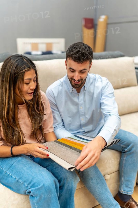 Young couple selecting upholstery swatches while sitting on a couch in a furniture showroom, making interior design decisions for their new living space