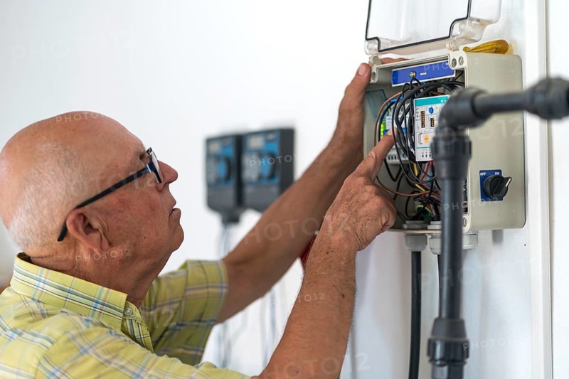 A senior man wearing glasses works attentively on an electrical panel, adjusting wires and using tools. The setting highlights his focused attention and technical skill