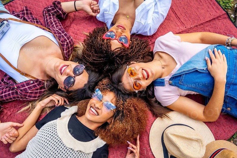 Four cheerful young women with sunglasses lying in circle on blanket enjoying sunny day in park