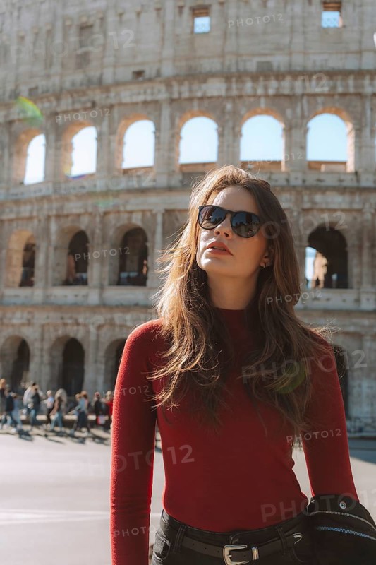 A stylish woman in sunglasses and a red top stands confidently before a historical European landmark on a sunny day, with tourists and ancient architecture in the background
