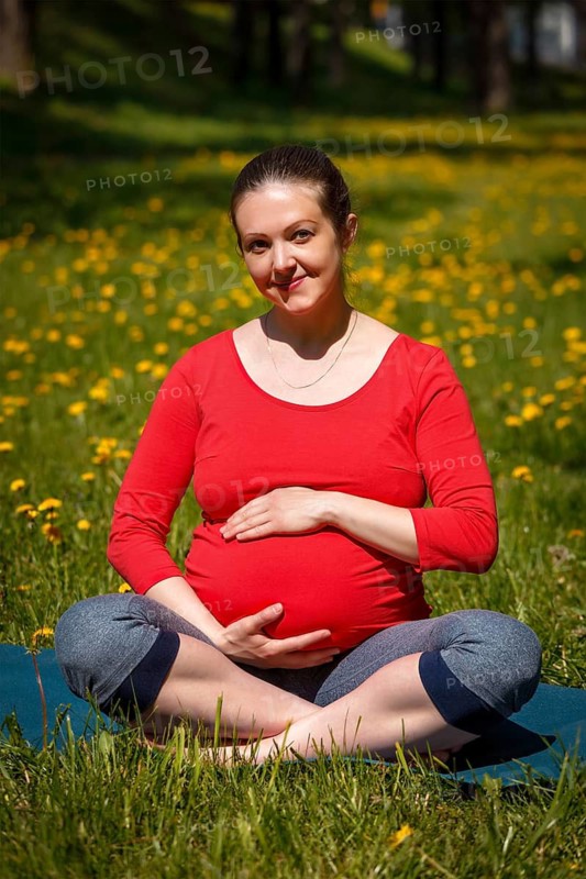 Pregnancy yoga exercise, pregnant smiling woman doing asana Sukhasana easy yoga pose holding her abdomen outdoors on grass lawn with dandelions in summer
