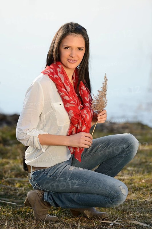 A woman in casual attire kneeling outdoors, holding a plant, and wearing a red scarf and white shirt, Bavaria