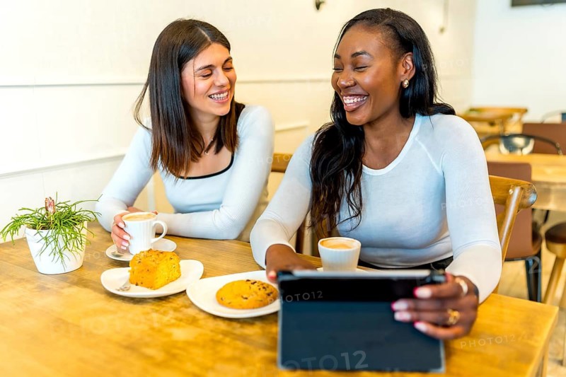 Two female friends are enjoying coffee and pastries while using a tablet in a cafe, laughing together