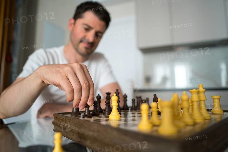 A mature man sits at a table, deeply engaged in a game of chess at home. He thoughtfully considers his next move, creating a moment of quiet concentration and strategy