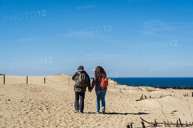 A couple of backpacker tourists travel through the sand dunes on the shores of the Baltic Sea, A couple walks hand in hand on a sandy path towards the ocean under a clear blue sky, Curonian Spit, Neringa, Lithuania