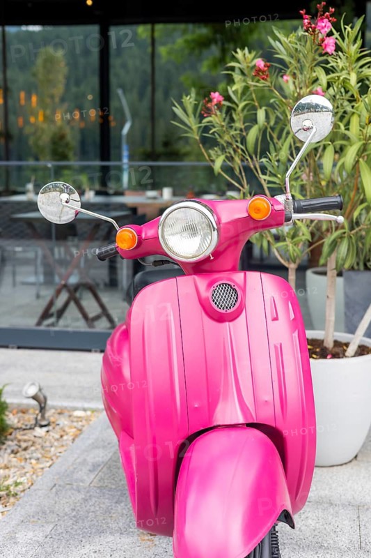 A vibrant pink scooter parked near a modern building with greenery. The scooter features a classic design with chrome mirrors and a shiny finish