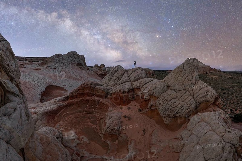 A distant, unrecognizable hiker stands atop a unique rock formation in Utah, USA, under a star-filled sky, showcasing the beauty of an outdoor adventure at night