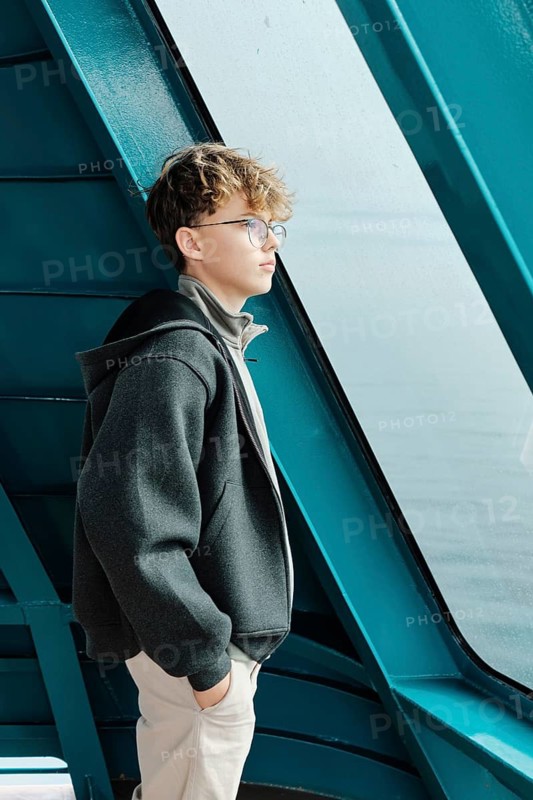 Teenage boy with curly hair and glasses gazes out the window at a ski resort. Dressed in a warm hoodie and jacket, he enjoys the peaceful winter landscape and the adventure of the mountains