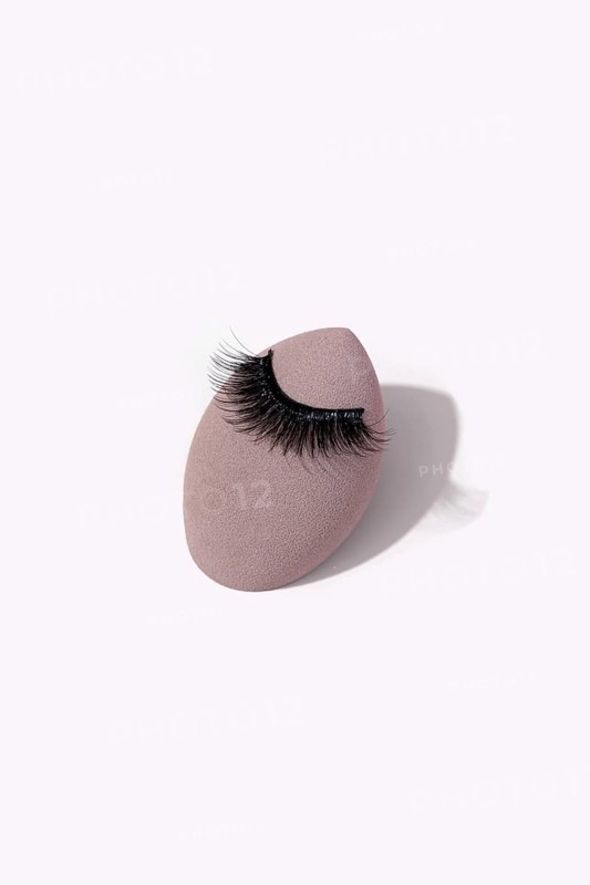Black false eyelashes is elegantly placed on a makeup sponge against a light background. This setup represents beauty essentials and minimalist aesthetics