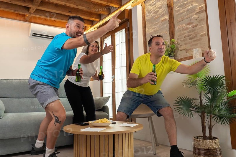 Friends standing up and celebrating a goal while watching a soccer match on TV at home. Excited gestures, raised arms, beer, snacks and shared joy indoors