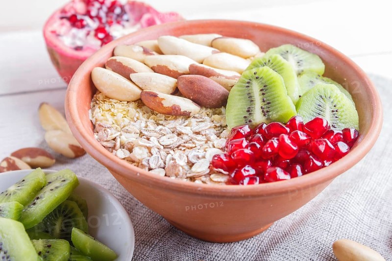 A plate with muesli, kiwi, pomegranate, Brazil nuts on a white wooden background. close up