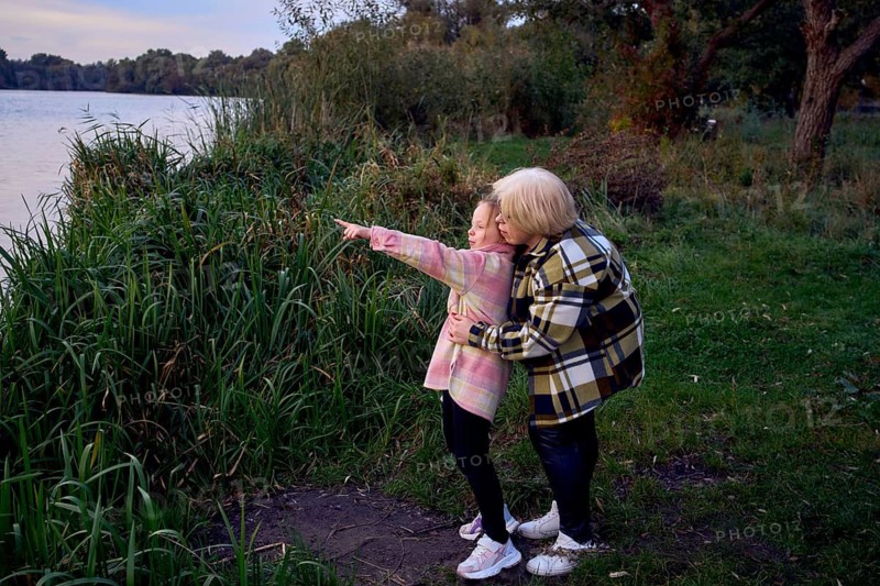 A mother and daughter share a peaceful moment by a river. The girl points toward the horizon, surrounded by lush greenery, symbolizing family bonds and exploration