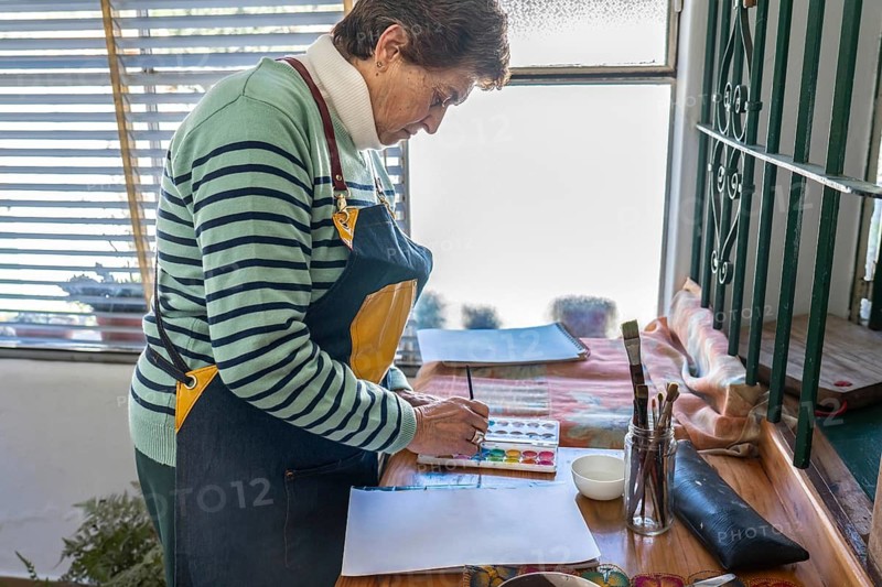 Older woman in denim apron stands over a wooden desk organizing watercolor supplies — palette, brushes, and paper — beside a barred window with natural light