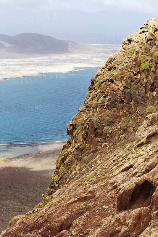 Rocky cliffs overlook the blue sea, under a wide sky, La Graciosa in the distance in the background, Lanzarote, Canary Islands, Spain