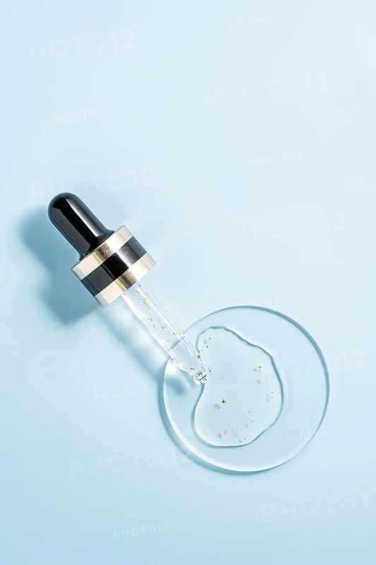 Close-up of a serum dropper with transparent cosmetic liquid placed on a light blue background, showcasing purity and skincare beauty with gold flecks visible in the serum
