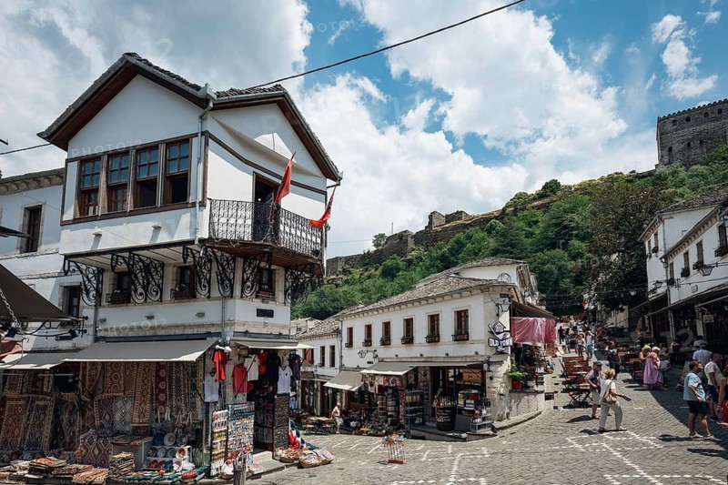 GJIROKASTER, ALBANIA - JUNE 30, 2026: Traditional Albanian street in Gjirokastër with white Ottoman-style houses and market stalls, real lifestyle travel illustration under summer sky