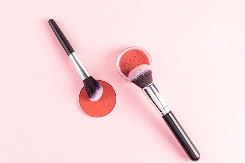 A stylish arrangement of makeup brushes with bristles dipped in a warm-hued compact blush, set against a soft pink backdrop. Perfect for beauty and cosmetics themes