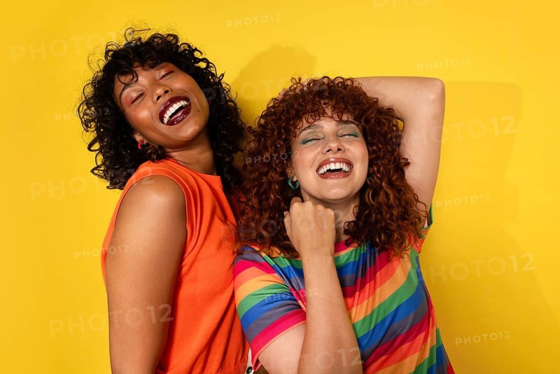 Two friends joyfully laugh together against a vibrant yellow background, their colorful outfits reflecting their cheerful energy and the warmth of their connection