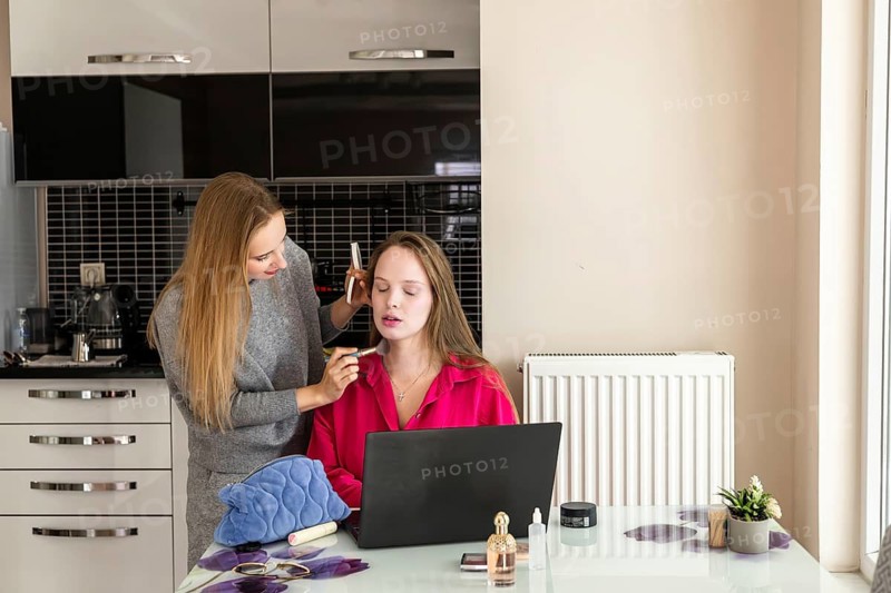 A young Caucasian woman with long blond hair applies makeup to another young woman with long brown hair at a modern kitchen table. A laptop and beauty products are visible