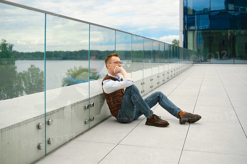 Middle-aged man sits on floor with back against glass railing. He looks thoughtful while gazing at river. Surroundings include modern buildings and cloudy sky. Time appears to be daytime