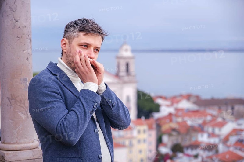 Man musician in a blue blazer and white sweater playing blues on a harmonica with eyes closed, standing outdoors in Lisbon city street, Portugal