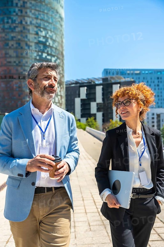 Two business professionals stroll through the city, chatting over coffee and a laptop, portraying modern urban corporate life, collaboration and confident, casual teamwork