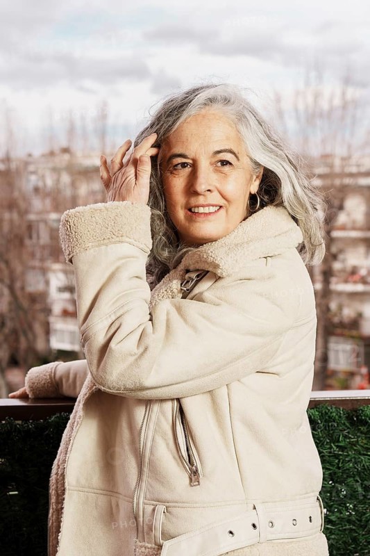 Smiling elderly woman with white hair stands on her balcony, wearing a cozy jacket. The background shows an overcast cityscape, enhancing the peaceful atmosphere