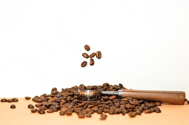 A wooden handled scoop rests among a pile of coffee beans, with several beans captured mid air, creating a dynamic display. The background is a simple white, emphasizing the beans