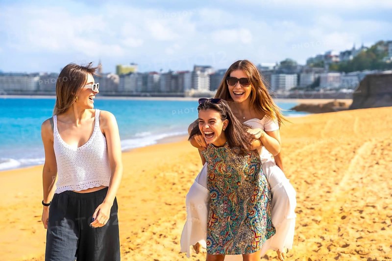 Three friends spending summer vacation together, two women sharing a joyful piggyback ride on the sandy beach while laughing and smiling under a sunny sky in san sebastian