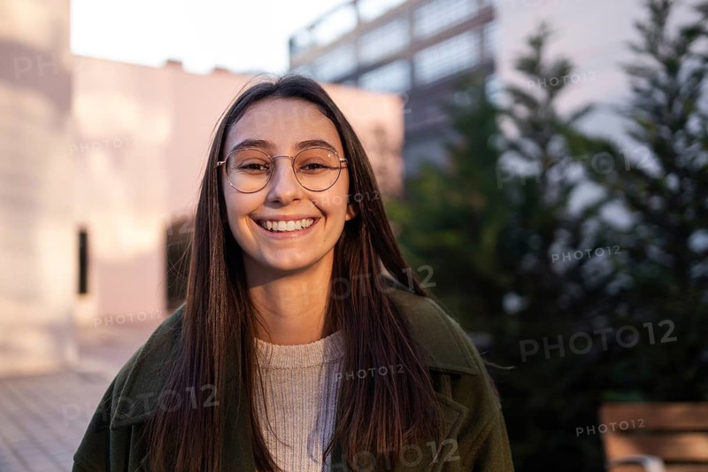 A woman with long brown hair and glasses smiles warmly outdoors She's wearing a green coat and beige sweater, with trees and a modern building in the background