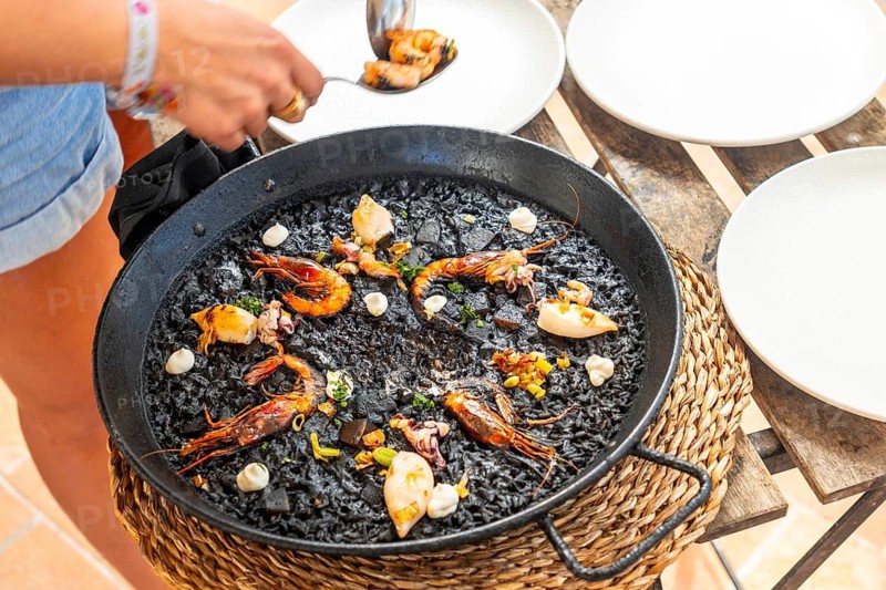 Chef serving a traditional black rice paella with shrimps and seafood, using a spoon, directly from the pan, in mallorca