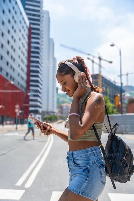 Young woman with headphones and phone feeling happy while navigating a bright summer day street, urban lifestyle and mobile technology connecting people with their favorite sounds
