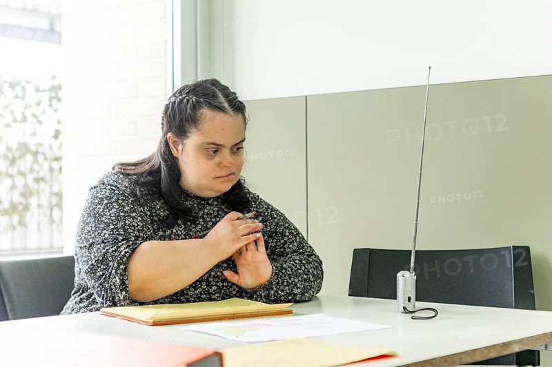 A woman is sitting at a table with documents in a mental health residence, looking contemplative, she is attentive while listening to the radio