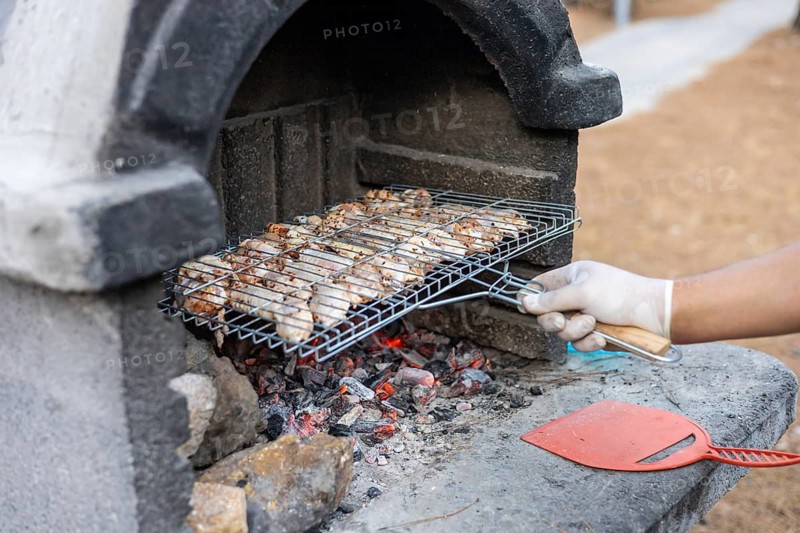 A hand with a glove holds a grill rack with cooked meat over hot coals. The scene is set in a park with trees in the background