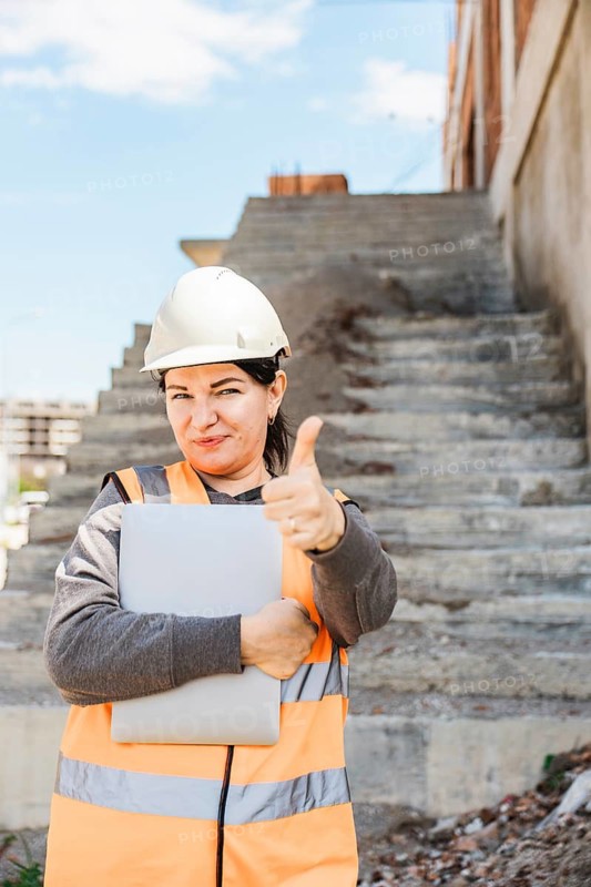 A young Hispanic woman in a hard hat and safety vest stands on construction stairs, giving a thumbs up while holding a laptop