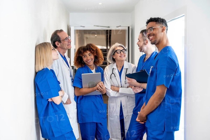 Happy multiethnic medical team of doctors and nurses in scrubs and lab coats standing in a clinic corridor, smiling and exchanging ideas while using a digital tablet