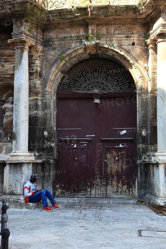Palermo, main entrance of a morbid house at the square Piazza Bologna in the old town, in front of it sits a young African with a mobile phone, Sicily, Italy