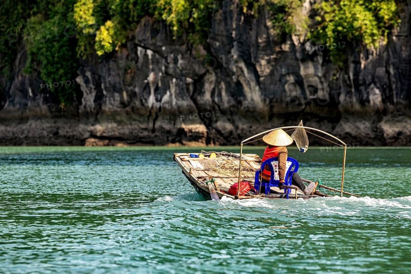 A dugout canoe with a seated person glides silently through clear, green water against a forested background, fishermen and fishing boats in Halong Bay, Vietnam