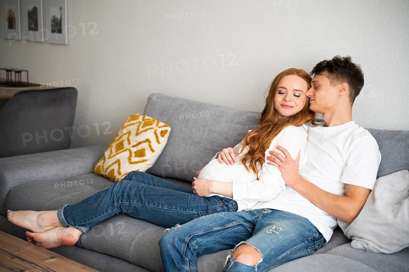 A young couple, joyfully expecting a baby, relaxes on a comfortable sofa in a cozy living room. They tenderly embrace, radiating happiness and anticipation for their new family member