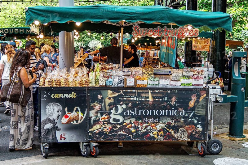 A mobile Gastronomica market cart advertising cannoli and sweet truffles displays trays of Italian confectionery beneath a striped green awning, with shoppers browsing the counter along one of the covered lanes of Borough Market in Southwark, London, England, United Kingdom