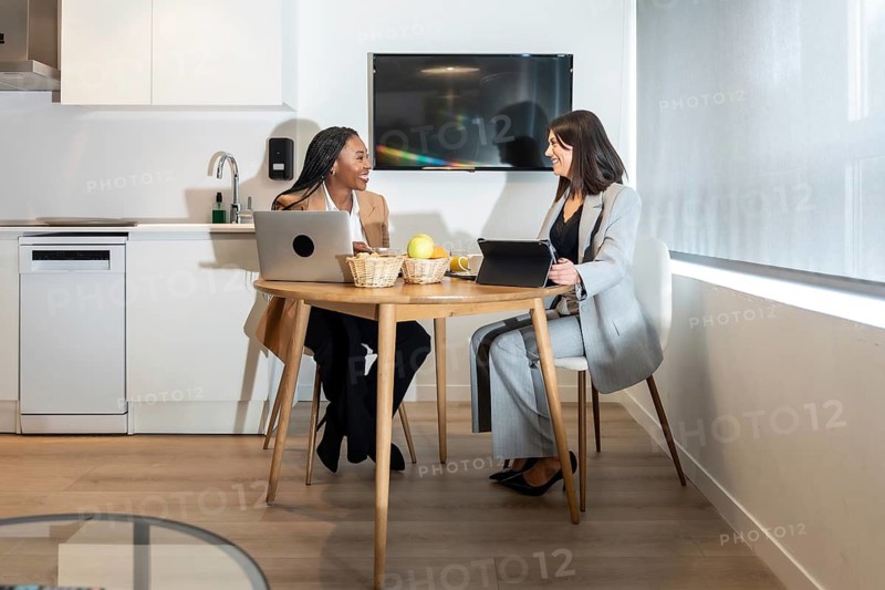 Two businesswomen are sitting at a table in a hotel room, working on their laptops and tablets while having a discussion