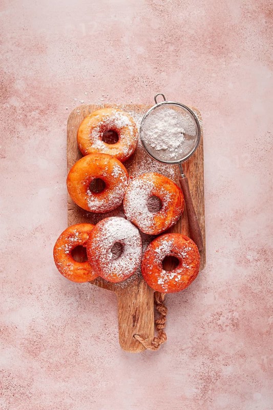 A group of baked donuts sits on a wooden cutting board. Powdered sugar is dusted on the donuts. This scene occurs in a kitchen setting, showcasing baked treats